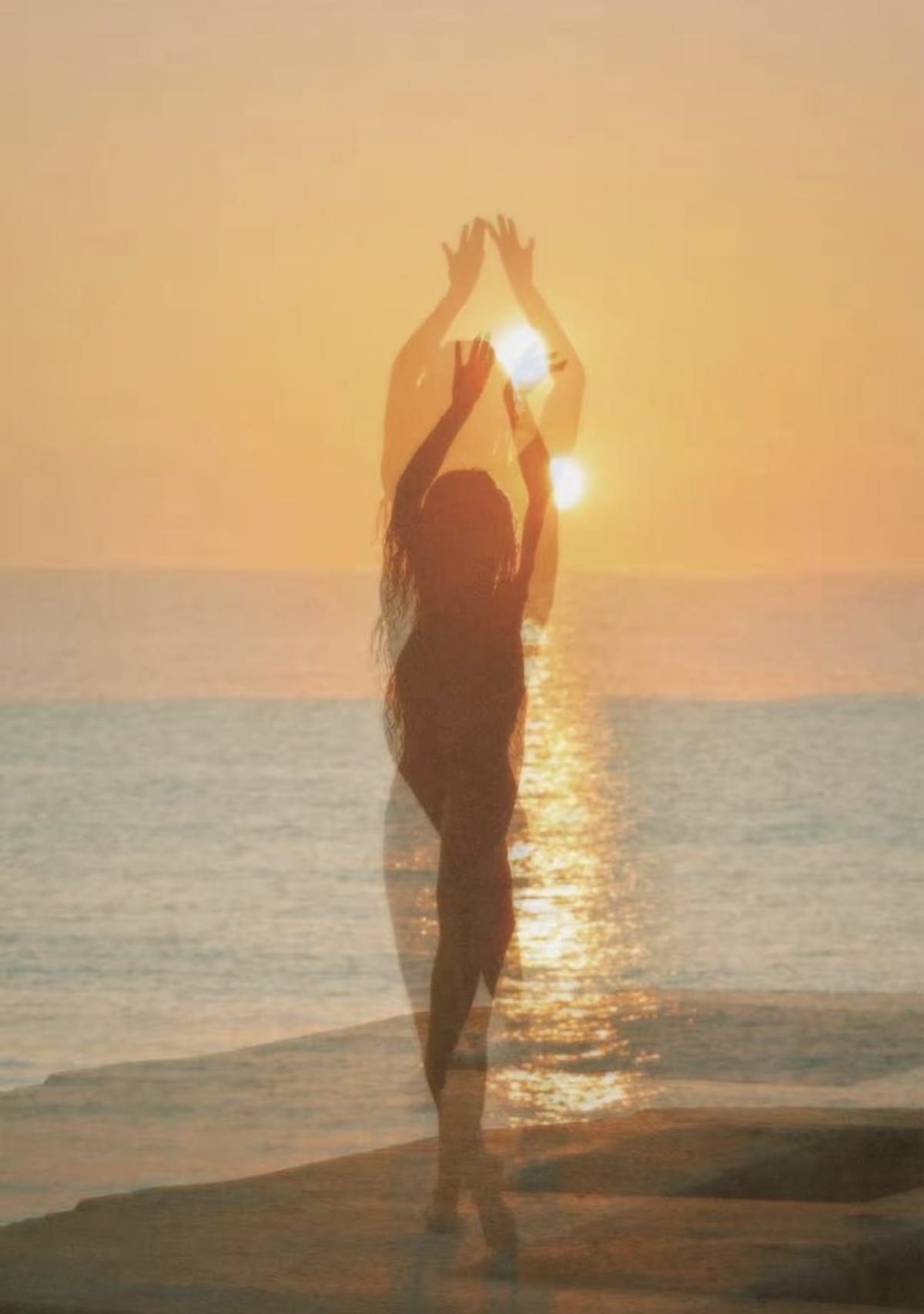 A woman silhouetted at the shoreline reaching toward a low sun over the ocean