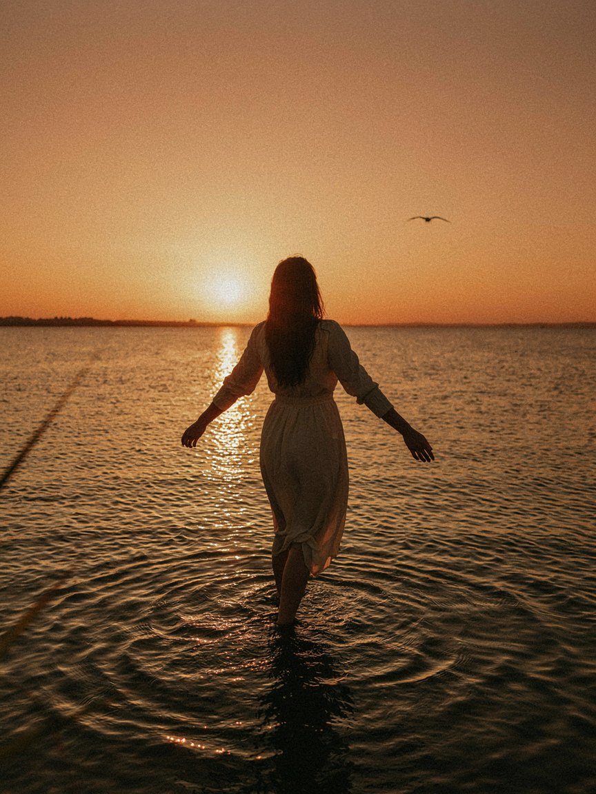 A woman standing in shallow water at sunset, arms open, facing the low sun