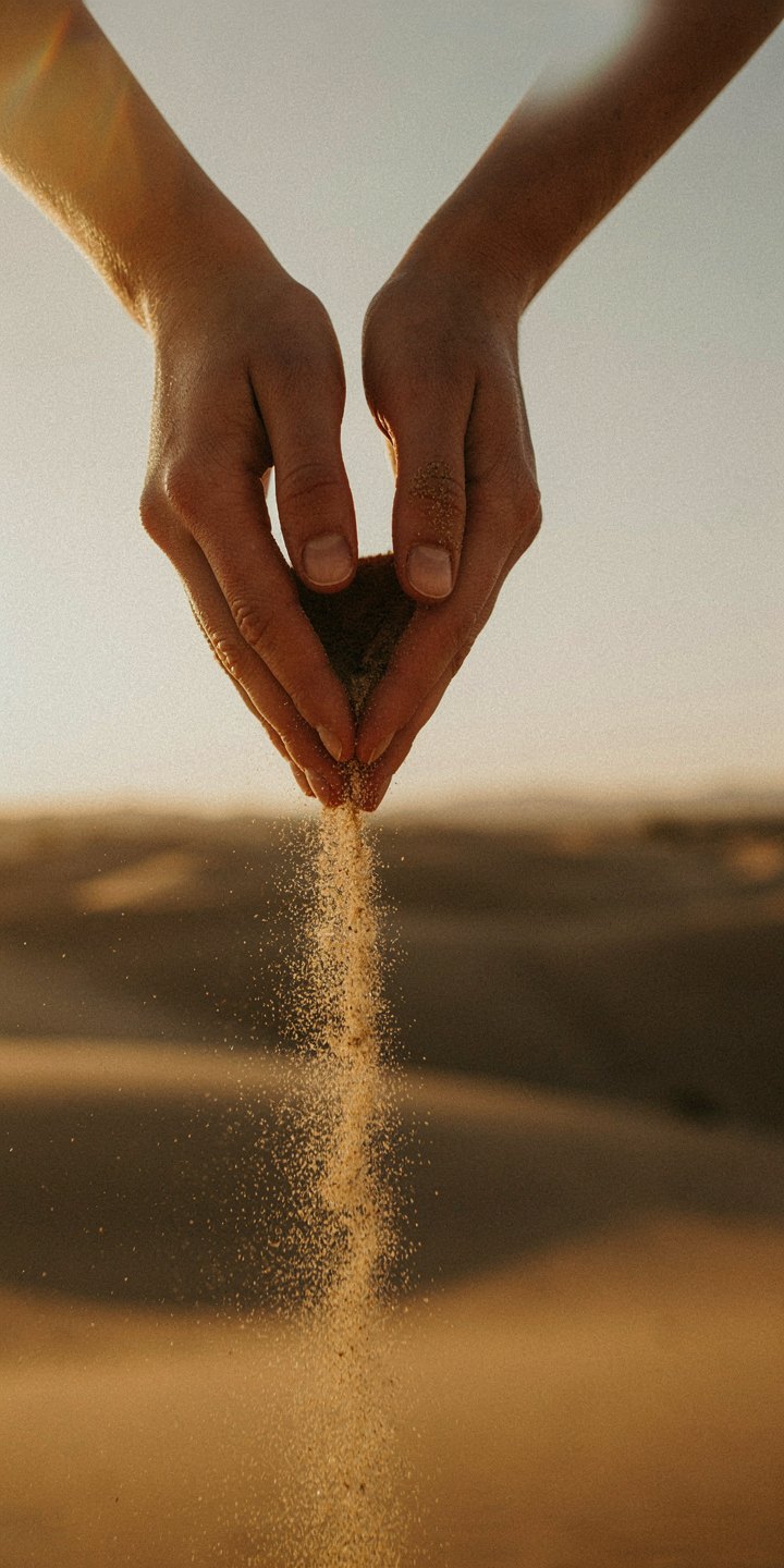 Cupped hands letting fine sand pour in a thin stream over warm dunes at sunset
